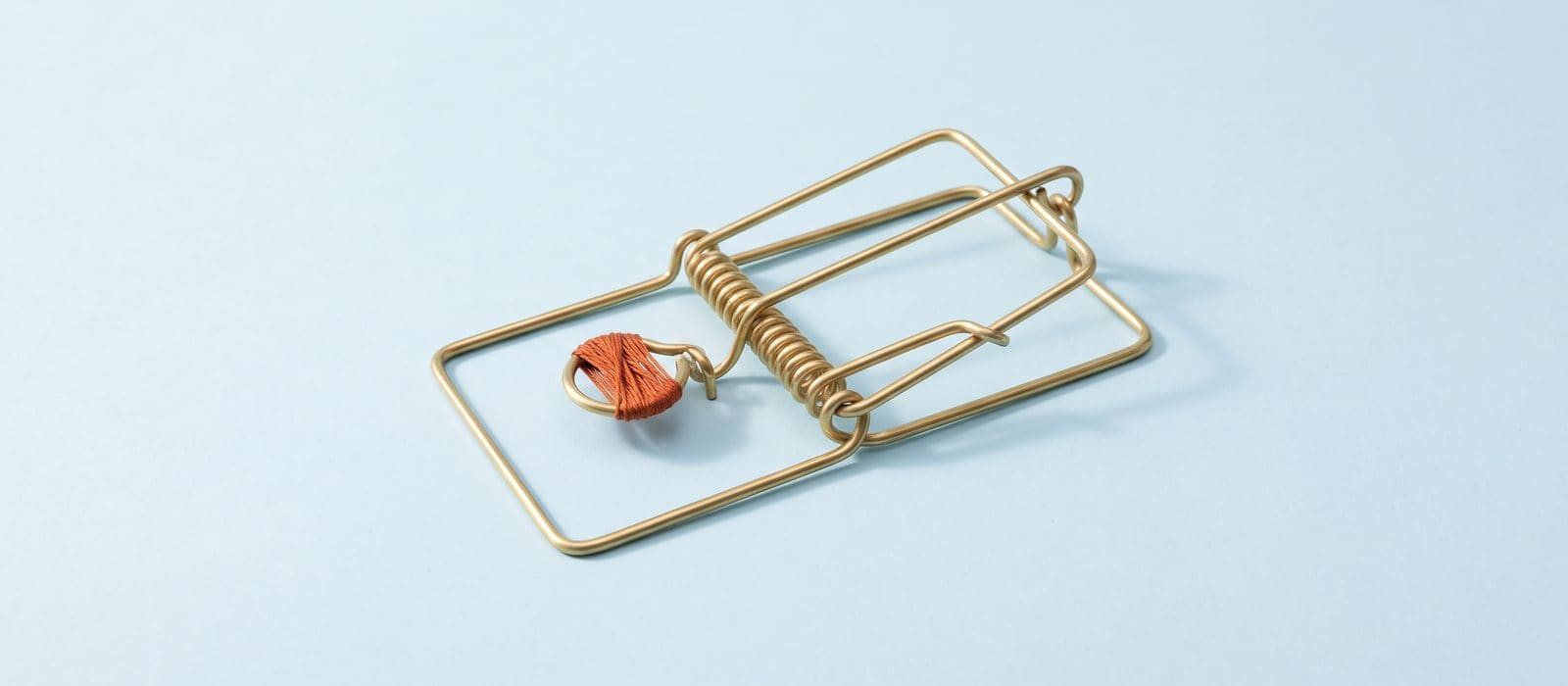 Brass wire sculpture of an empty mousetrap, its bait pedal wrapped in burnt-orange thread, on a pale-blue backdrop