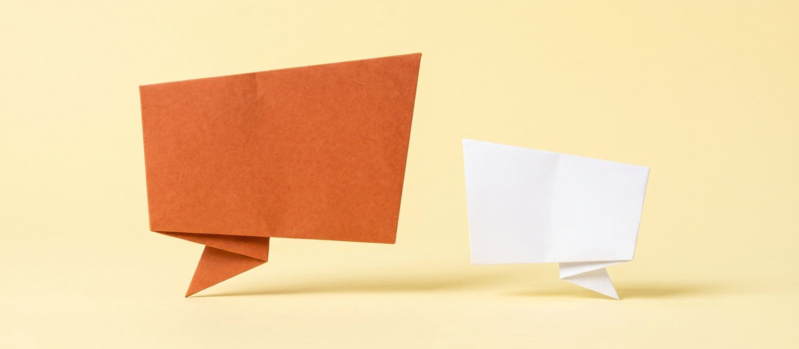 Two blank origami speech bubbles, one burnt orange and one white, on a pale yellow backdrop, representing the right to silence