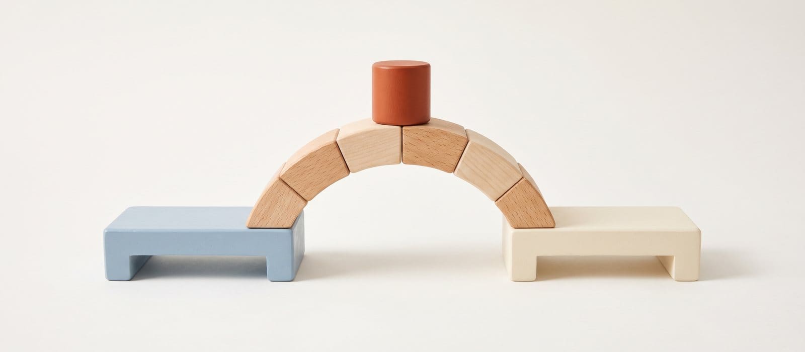 Wooden toy blocks forming an arched bridge between a soft blue platform and a cream platform, with an orange cylinder at the top
