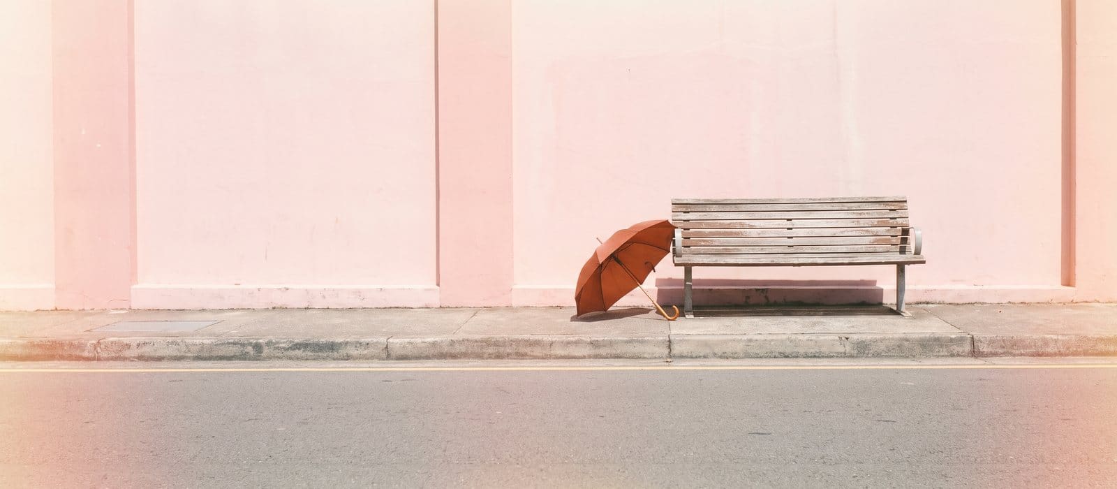 Instant-film photo of an empty wooden bench at a quiet kerbside pickup spot beside a pink wall, a burnt-orange umbrella leaning on it