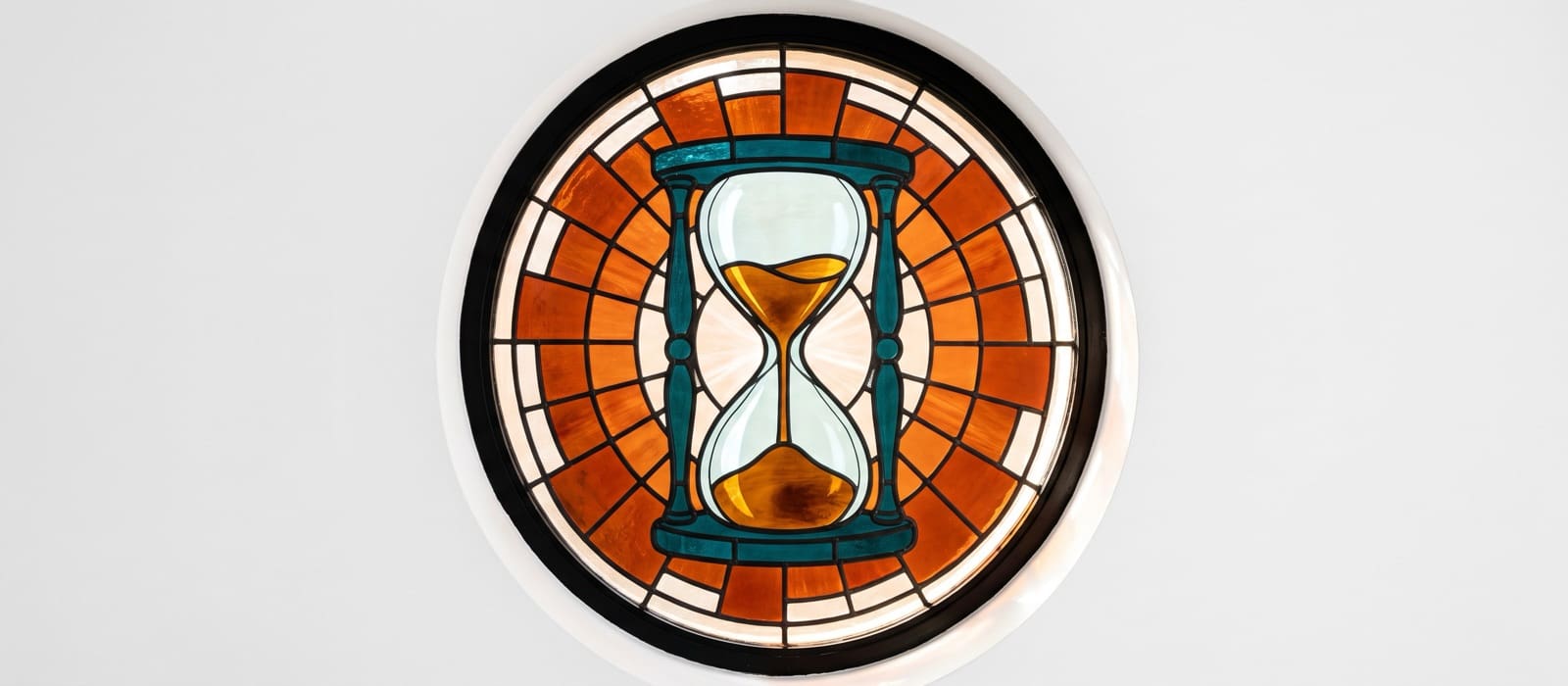 Round stained-glass window of an hourglass with amber sand running down, framed by burnt-orange panes