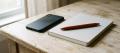 A phone lying face-down beside a closed notebook with an orange pen resting on it, on a wooden table by a window