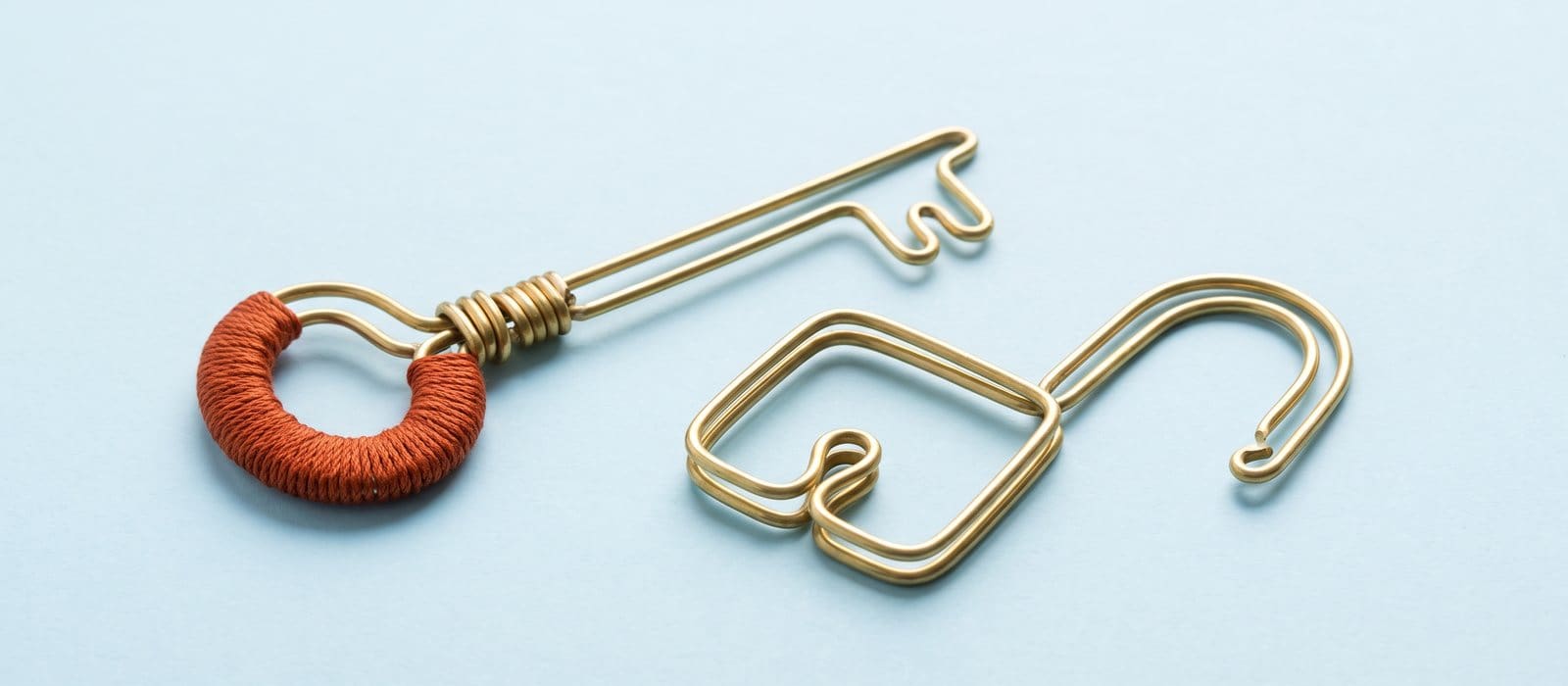 Brass wire sculpture of a key with a burnt-orange thread-wrapped bow beside an open padlock