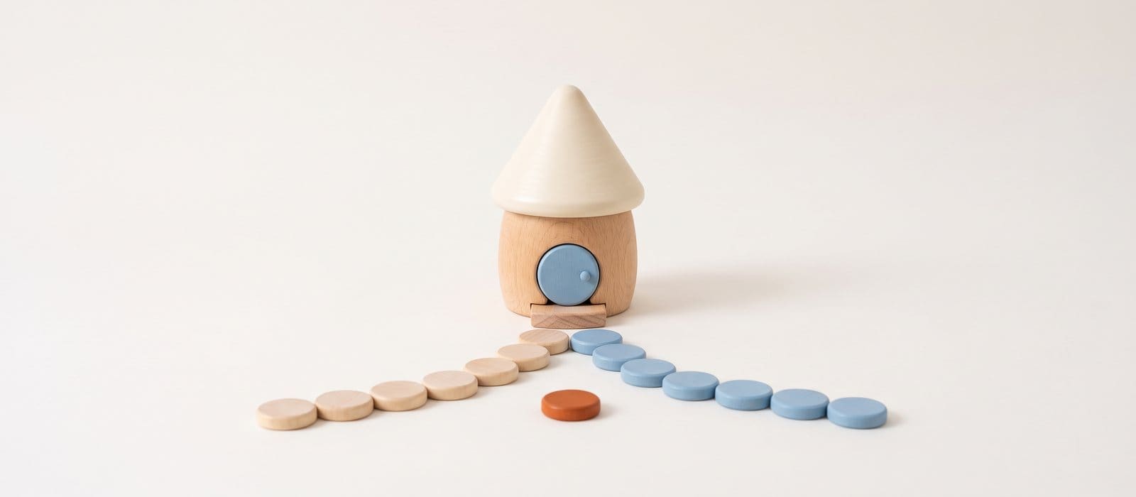 Turned-wood toy house with two paths of wooden discs leading away from its door, one natural maple and one soft blue, and an orange disc at the fork