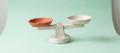 A handmade papier-mache balance scale with one burnt-orange pan and one plain pan, level on a pale mint background, representing the line between drug possession and drug trafficking