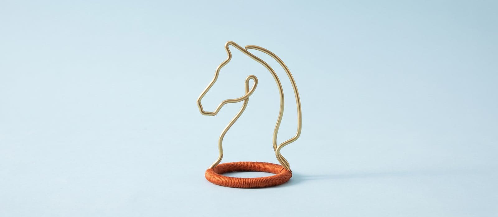 Brass wire sculpture of a chess knight on a base wrapped in burnt-orange thread, on a pale-blue backdrop
