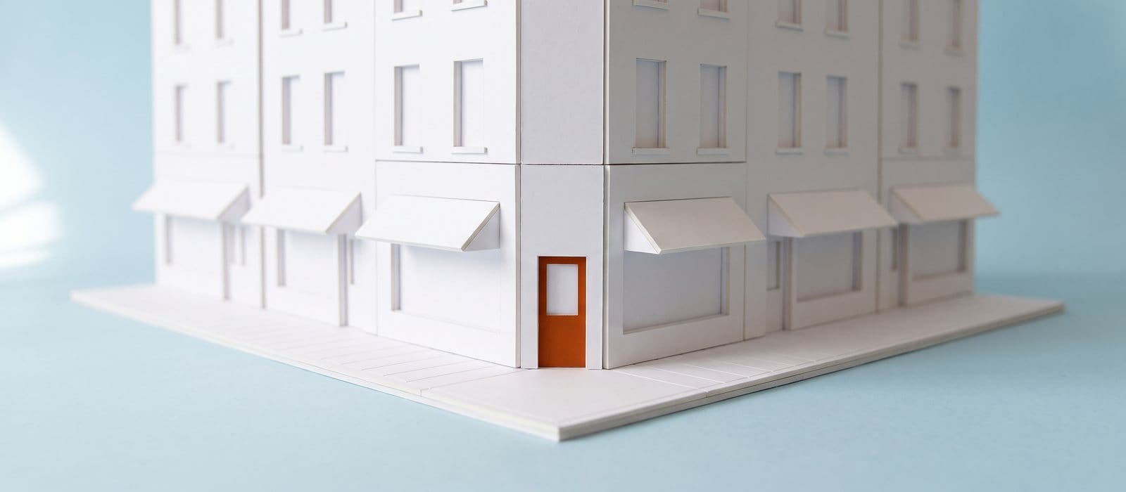 White card architectural model of a city street corner of shops with one burnt-orange shopfront door, on a pale blue backdrop