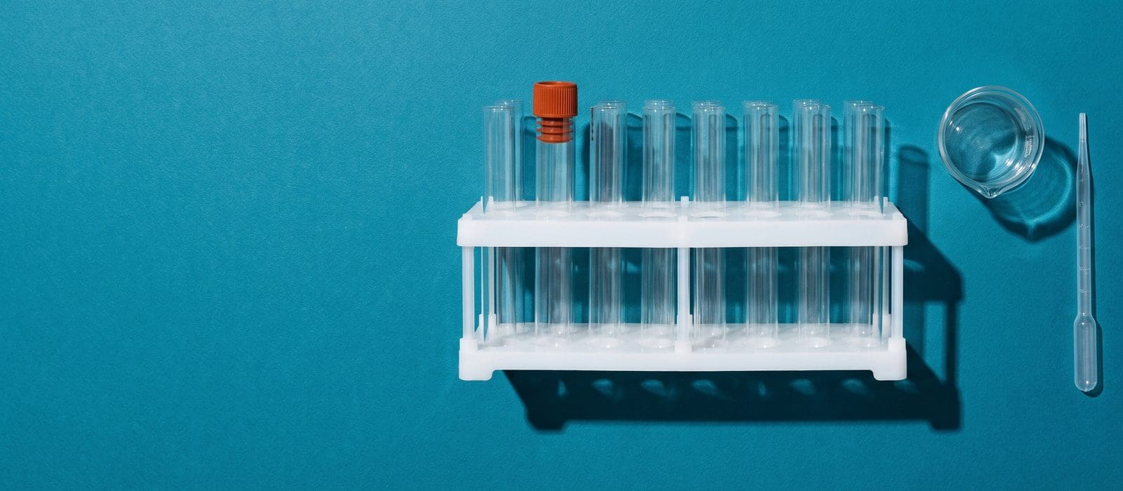A rack of empty test tubes with one orange-capped tube, a glass beaker and a pipette on deep teal paper, representing the lab analysis the Crown needs to prove a controlled substance