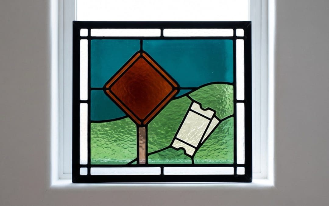 Stained-glass window of an orange diamond road sign and a pale paper ticket against a teal sky and green hills