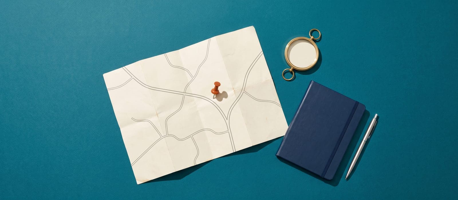 A cream road map with an orange push pin at a fork in the road, a brass compass and a navy notebook on deep teal paper, representing diversion away from prosecution