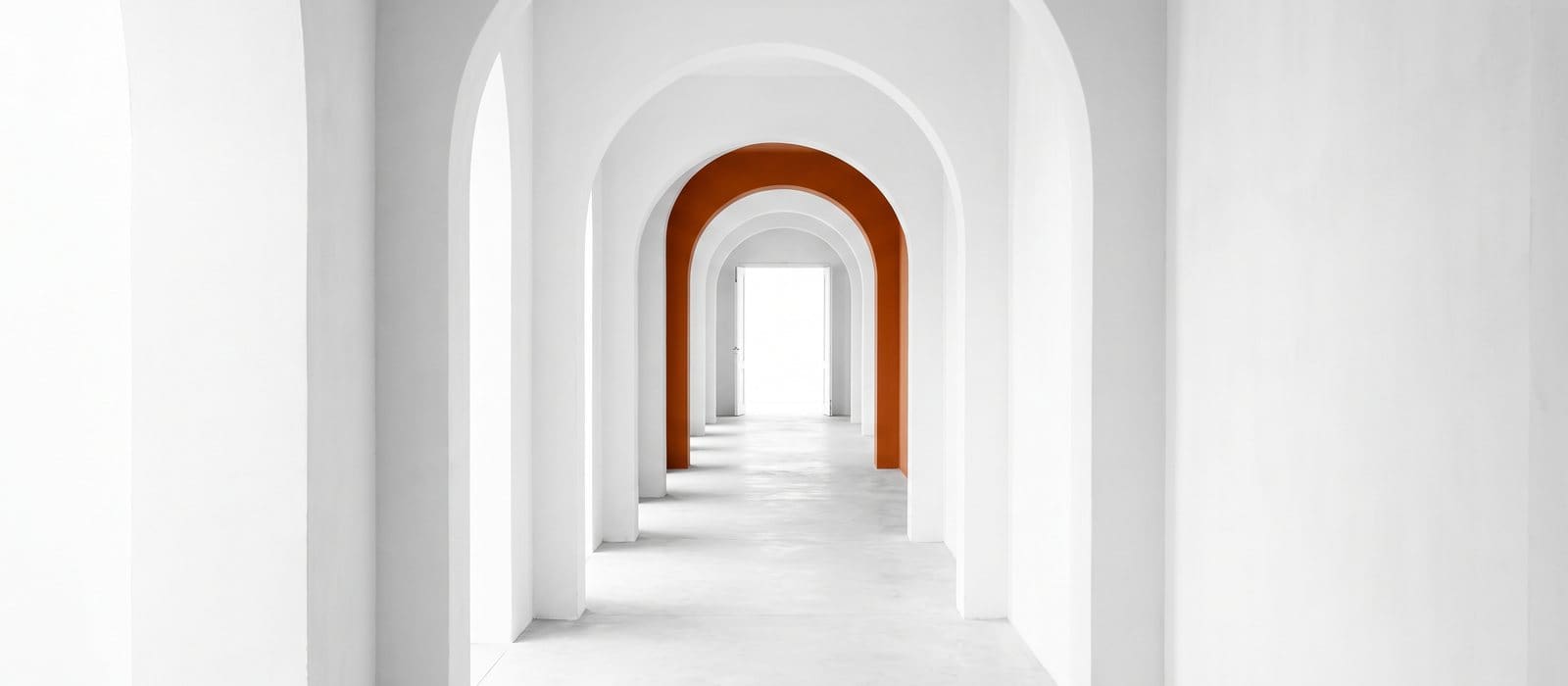 A long bright corridor of white arches, one arch in burnt orange, leading to an open doorway of daylight