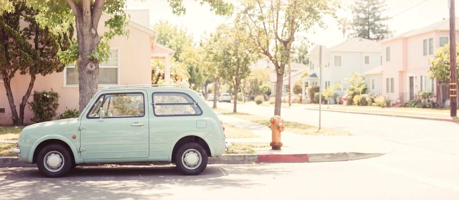 Instant-film photo of a mint-green car parked alone on a quiet sunny residential street beside an empty stretch of kerb and a burnt-orange fire hydrant