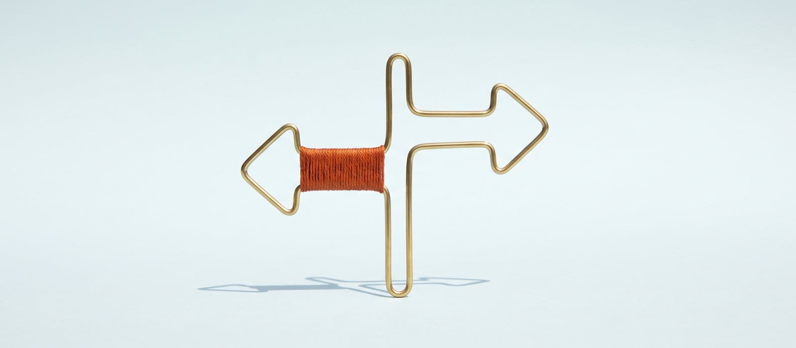Brass wire sculpture of a signpost with two arrows pointing opposite ways, one arm wrapped in burnt-orange thread