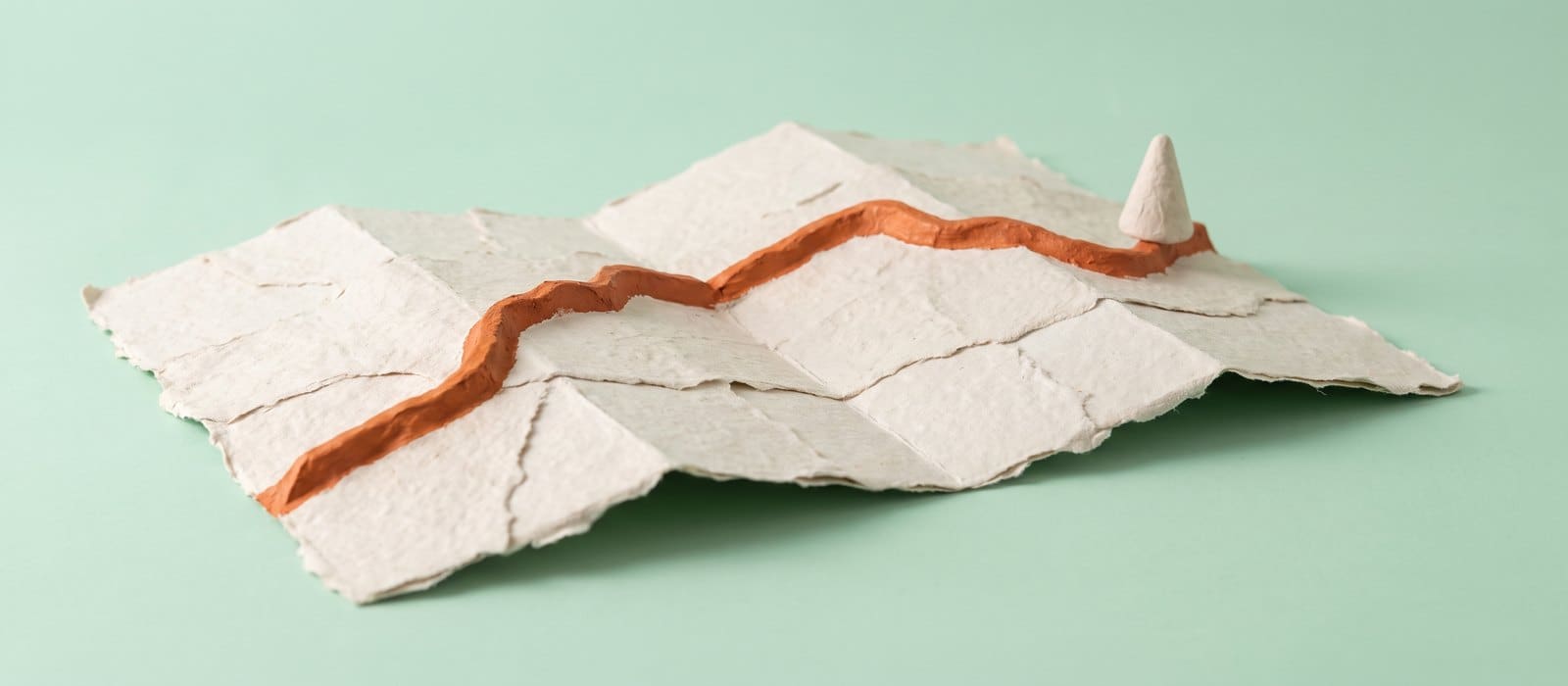 A handmade papier-mache folded road map with a raised burnt-orange route running across it and a small map pin, on a pale mint background, representing an importing or transport route