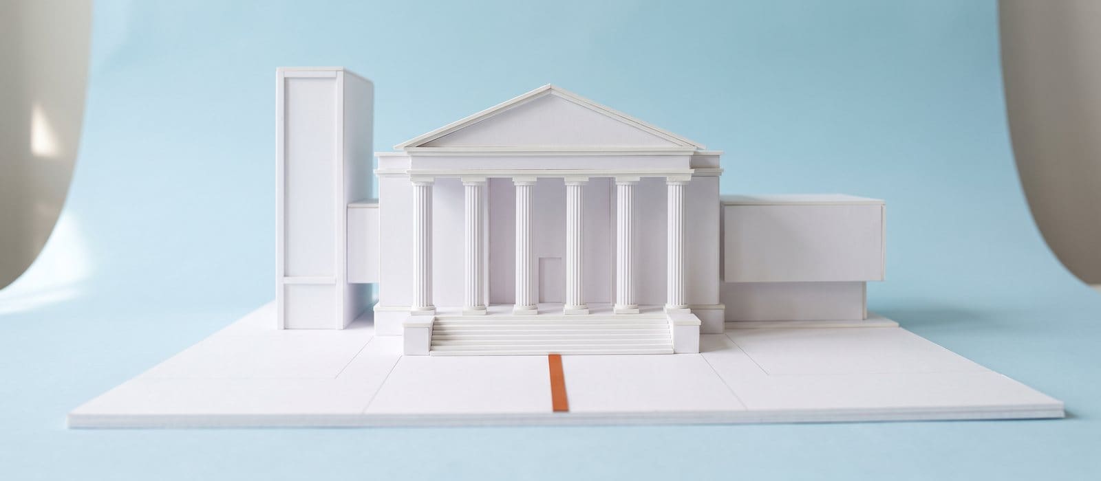White card scale model of a columned bank-style building with an orange line leading to its front steps, on a pale blue backdrop