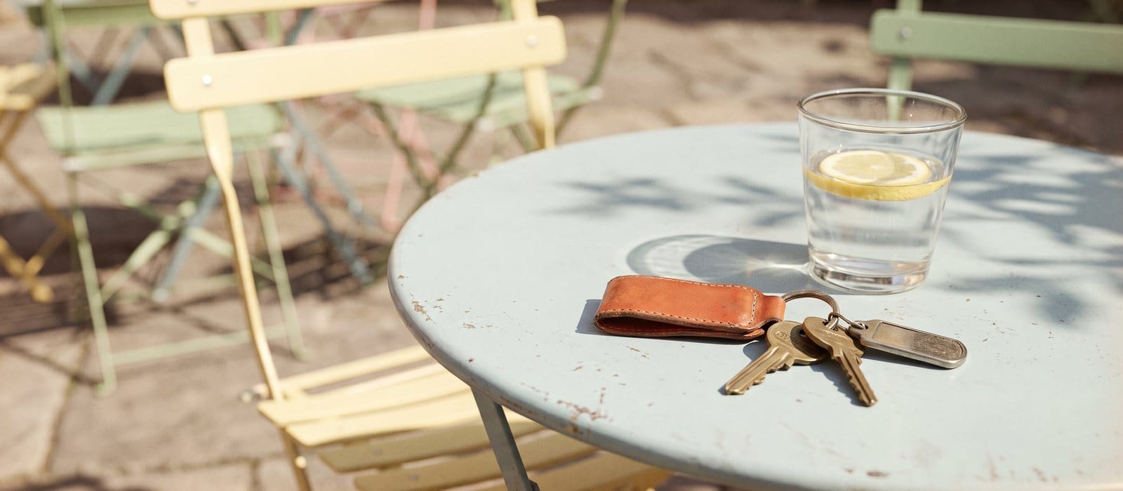 Instant-film photo of car keys on a burnt-orange leather fob beside a glass of water on a pale blue café table in sunshine