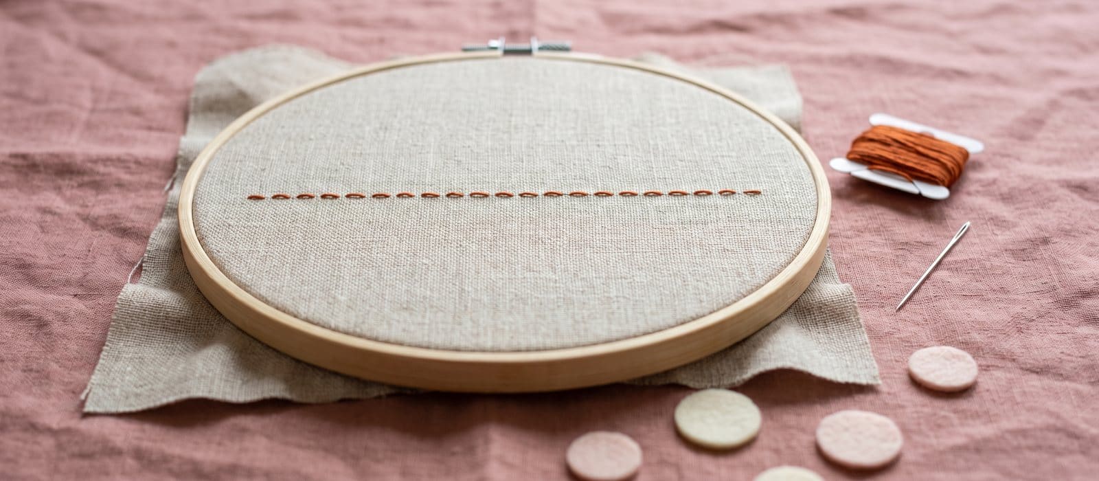 Wooden embroidery hoop holding linen with one straight line of burnt-orange running stitches, on dusty-rose linen