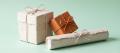Three handmade papier-mache parcels tied with string, one painted burnt orange, on a pale mint background, representing the transfer and delivery acts that make up trafficking