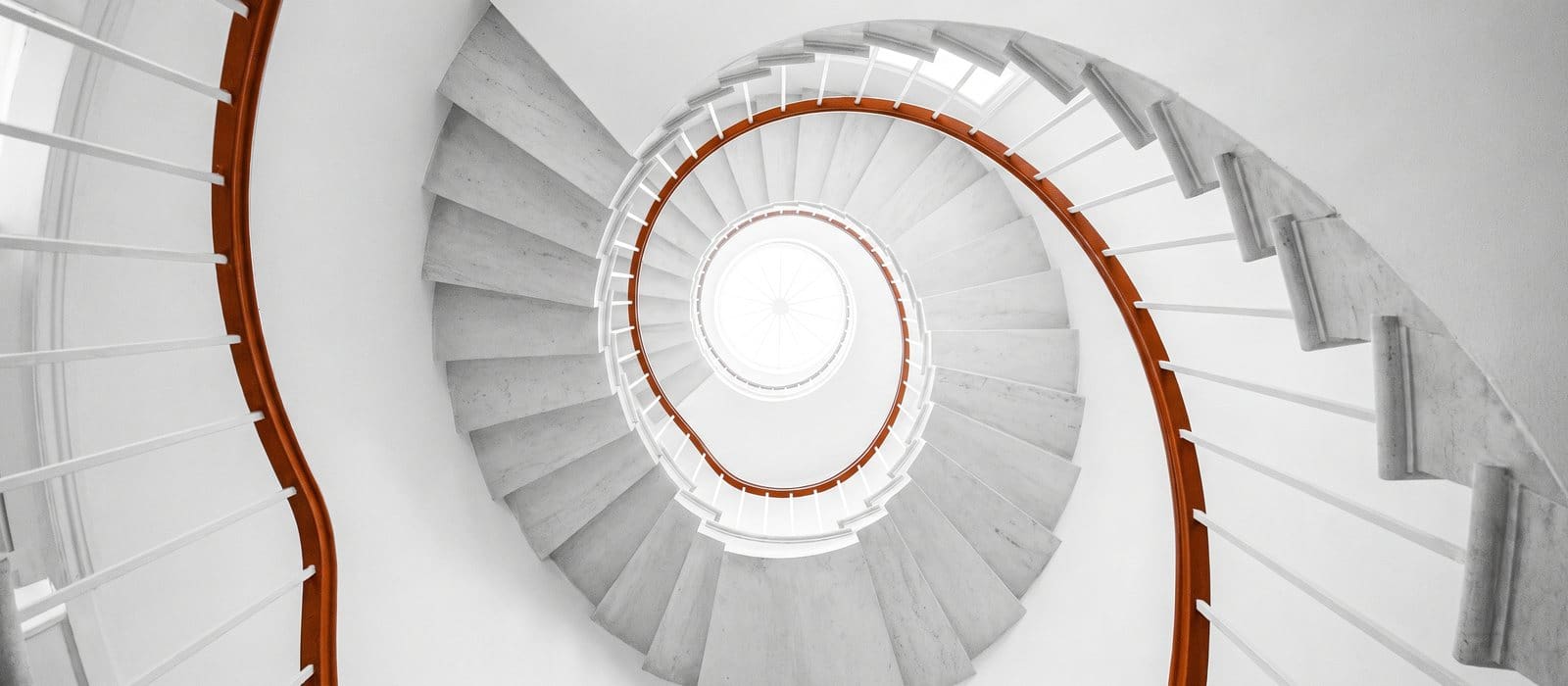 Looking up a bright white spiral staircase toward a skylight, its single handrail picked out in burnt orange