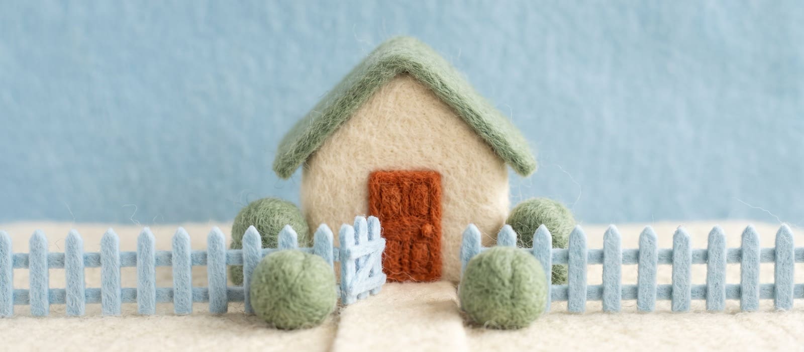 Needle-felted wool cottage with a sage roof, a burnt-orange front door and an open sky-blue garden gate