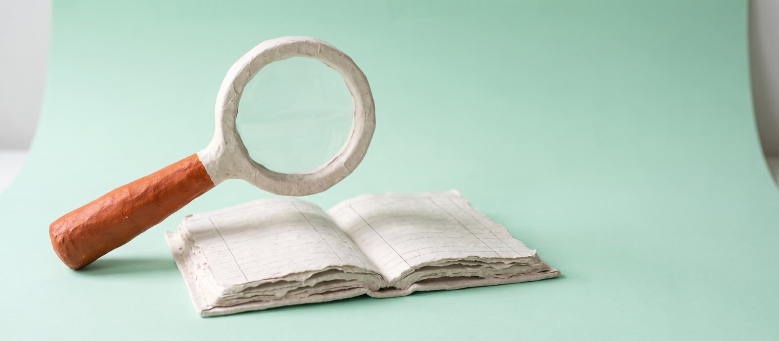 A handmade papier-mache magnifying glass with a burnt-orange handle hovering over an open blank ledger book on a pale mint background, representing a close review of the Crown's disclosure